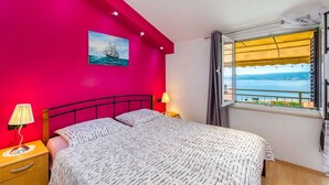 Quarto com cama, mesa de cabeceira, abajur, pintura e janela com vista para o mar.