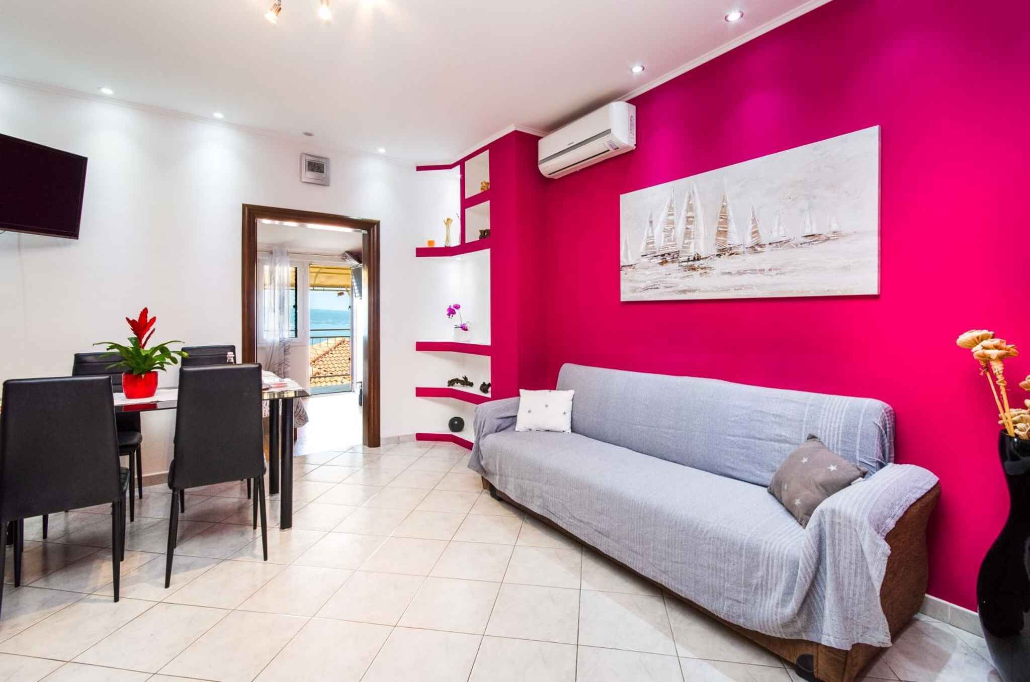 Un salon moderne avec un mur d’accent rose vif, un canapé gris, un coin repas avec une table et des chaises, et un tableau représentant des voiliers.
