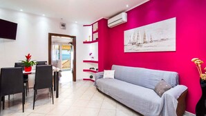 Uma sala de estar moderna com uma parede de destaque rosa vibrante, um sofá cinza, uma área de jantar com mesa e cadeiras, e um quadro com barcos à vela.