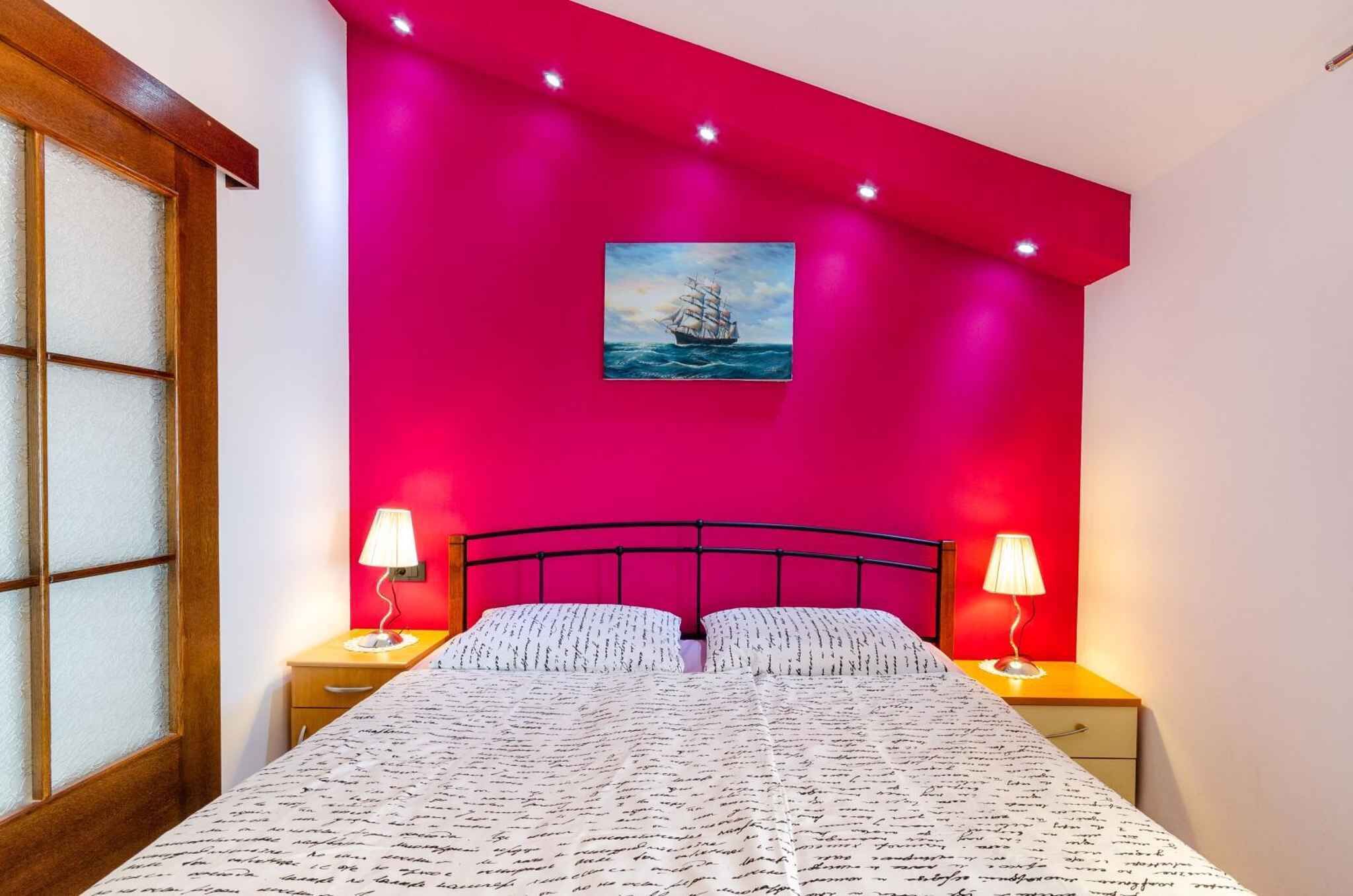 A bedroom with a bed, two bedside tables with lamps, a window with frosted glass, and a painting of a ship on the wall.