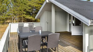 Un espacio comedor al aire libre con techo, una mesa, sillas, una terraza de madera y un edificio blanco con tejado gris.