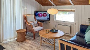 Una sala de estar acogedora con techo de madera, una silla de mimbre, una mesa redonda de madera y un televisor mostrando adornos coloridos.