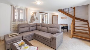 Un salon comprenant un canapé d’angle, une table basse avec un livre et un saladier de fruits, un escalier en bois et une table fixée au mur sur laquelle est posé un jeu de société.