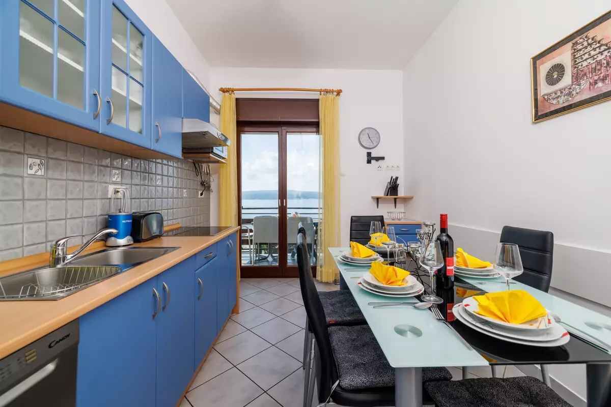 A modern kitchen with blue cabinets, a dining area with a table set for two, and a view through a glass door.