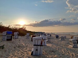 A beach with rows of white beach chairs and umbrellas, a pier in the background, and the sun setting over the horizon.