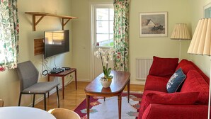 Ein Wohnzimmer mit einem roten Sofa, einem Holztisch, einem an der Wand montierten Fernseher, einem kleinen Tisch mit elektronischen Geräten und einem Fenster mit Vorhängen.