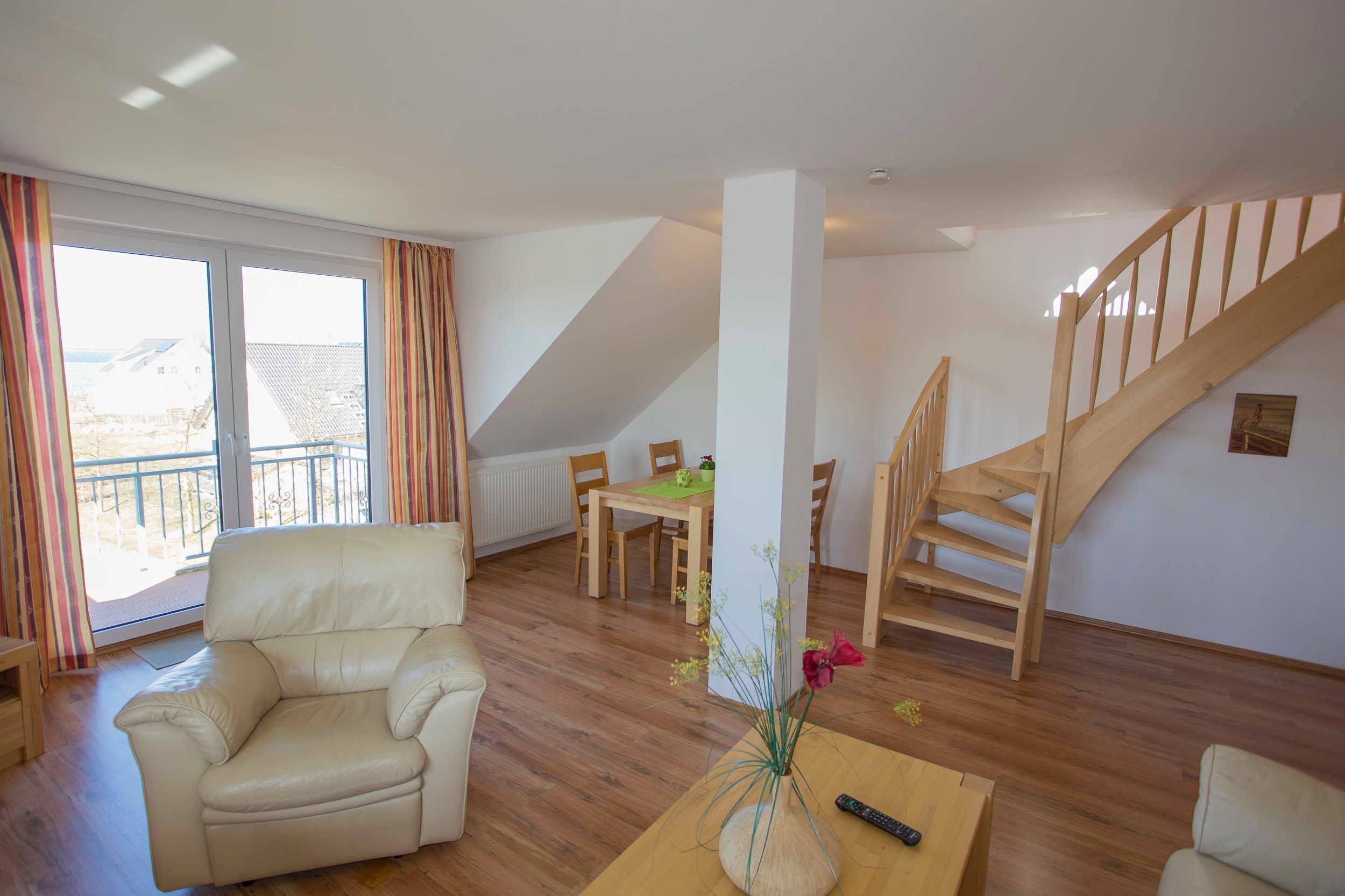 Un vaste salon comprenant un canapé en cuir, une table basse en bois et un coin repas avec une table et des chaises. Un escalier en bois est visible au fond.