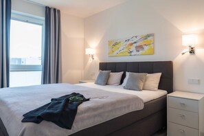 A modern hotel room with a large bed, a bedside table, a nightstand, a wall-mounted lamp, and a window with curtains.