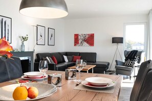 A modern living room with a dining area, featuring a wooden table set for two, a black leather sofa, and a large window providing natural light.