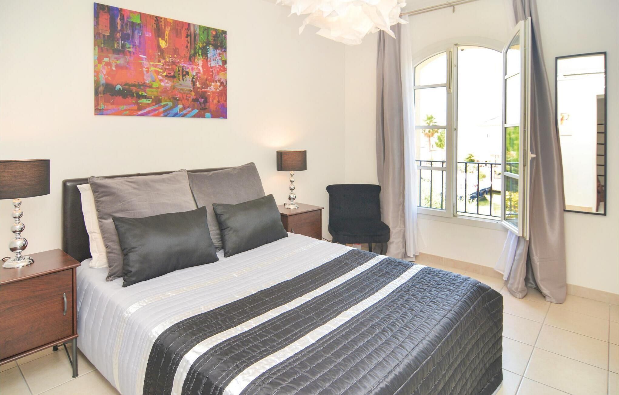 A bedroom with a bed, two bedside tables, a chair, a window with curtains, and a colorful painting on the wall.