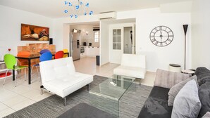 A modern living room with a white sofa, a glass coffee table, and colorful chairs. A kitchen is visible in the background. - Beautiful home in Aigues-Mortes with WiFi (Aigues-Mortes)