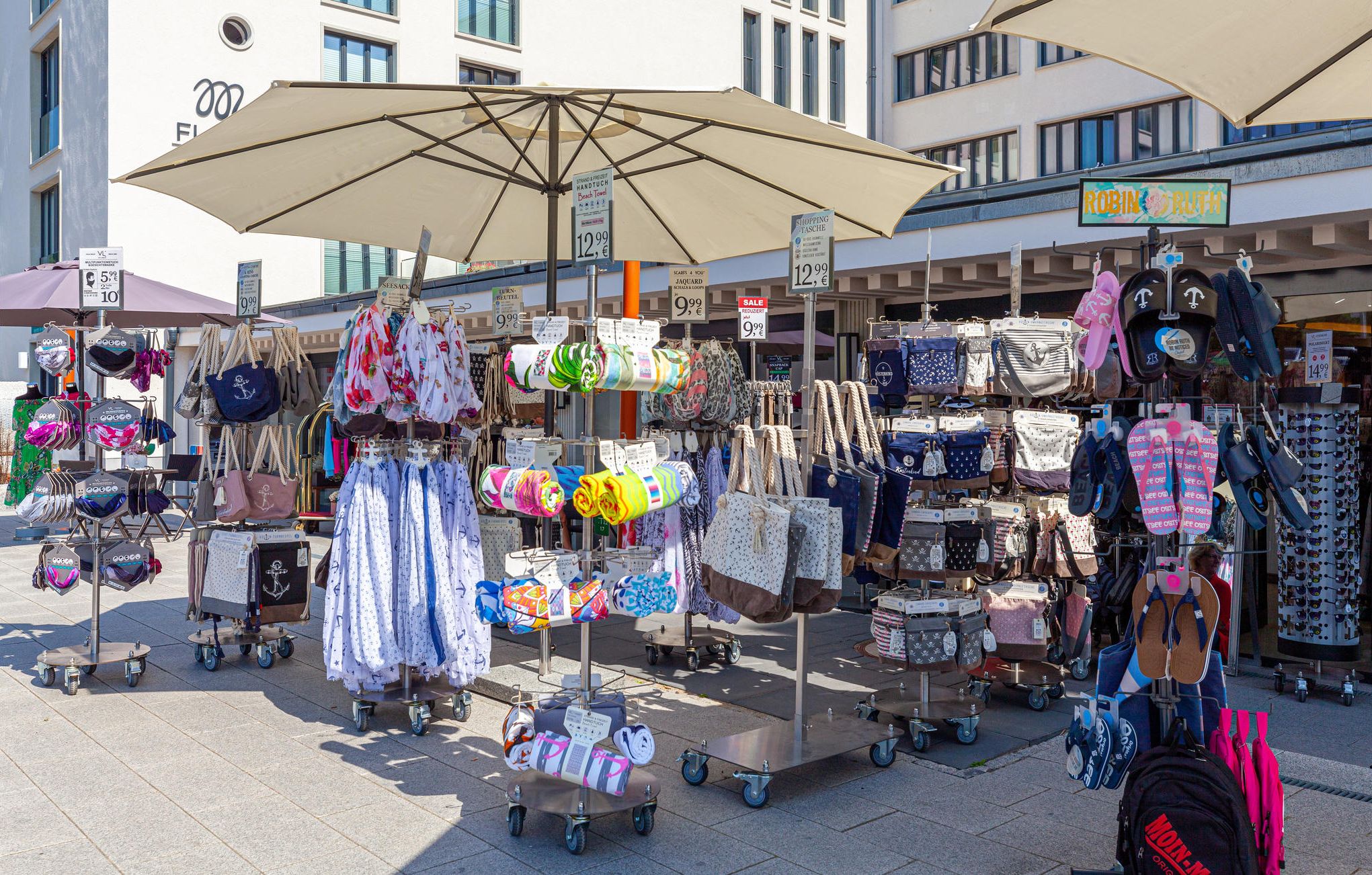 A market stall with various bags and accessories, including handbags and wallets, displayed on carts and under umbrellas.