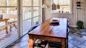 Une table à manger en bois, des chaises, une suspension et une fenêtre donnant sur une terrasse et des arbres.