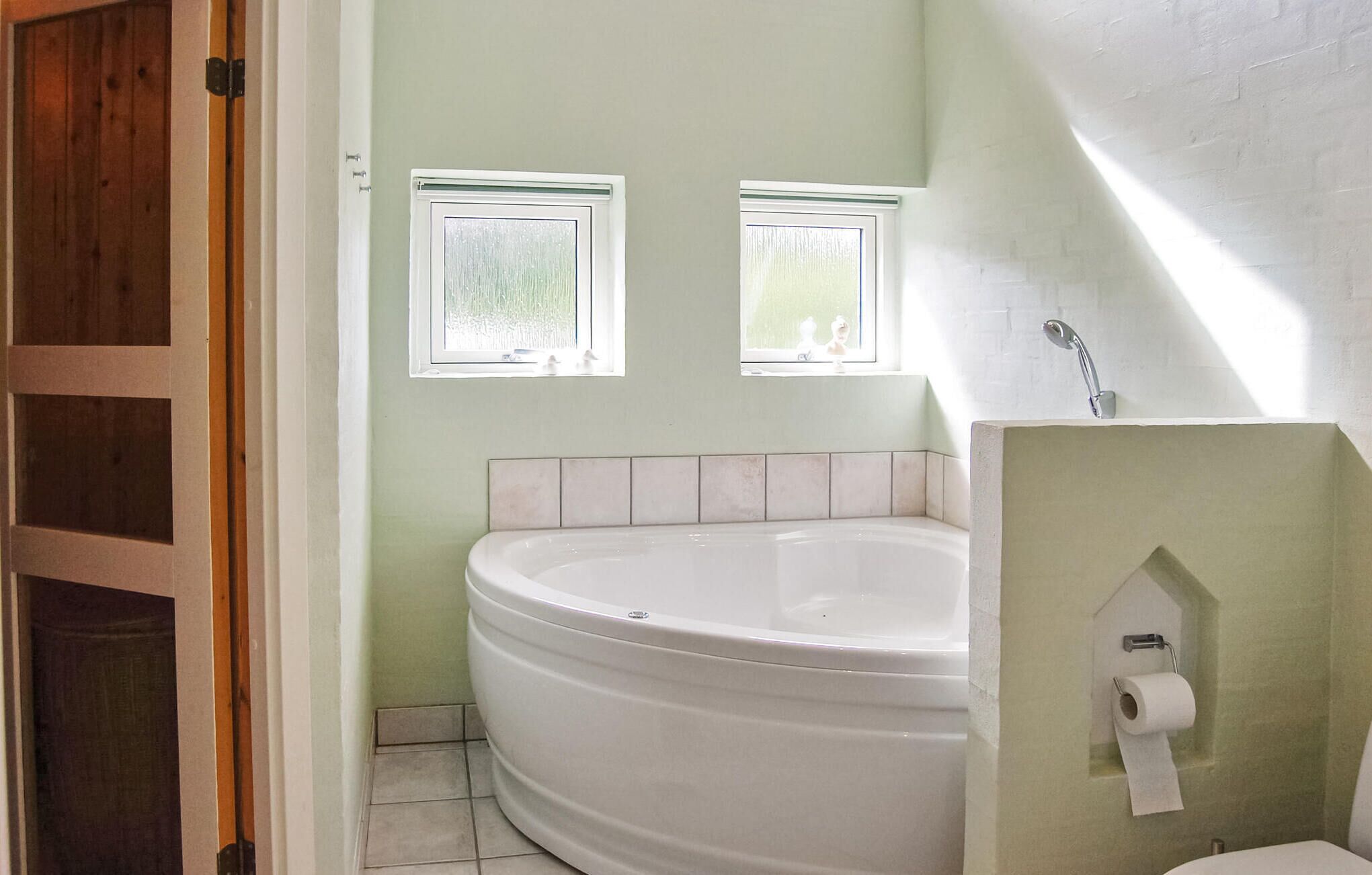 Um banheiro com banheira independente, duas janelas e vaso sanitário com suporte para toalhas de papel.