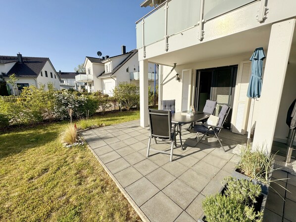 A patio with a table and chairs, surrounded by a garden and a white building with a covered entrance.