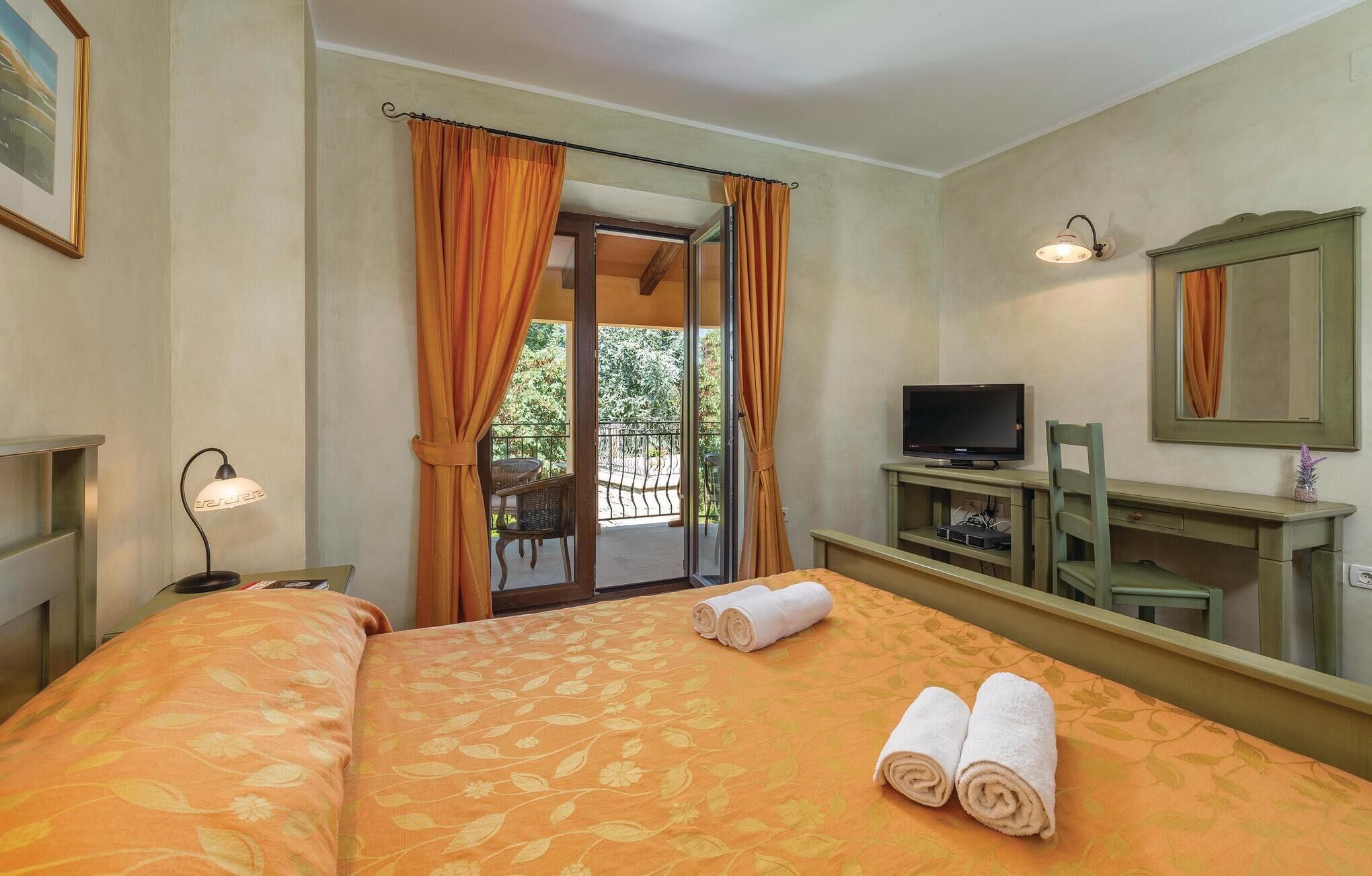 A hotel room with a bed, a TV, a desk, a chair, a lamp, and a balcony with curtains.