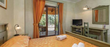A hotel room with a bed, a TV, a desk, a chair, a lamp, and a balcony with curtains.