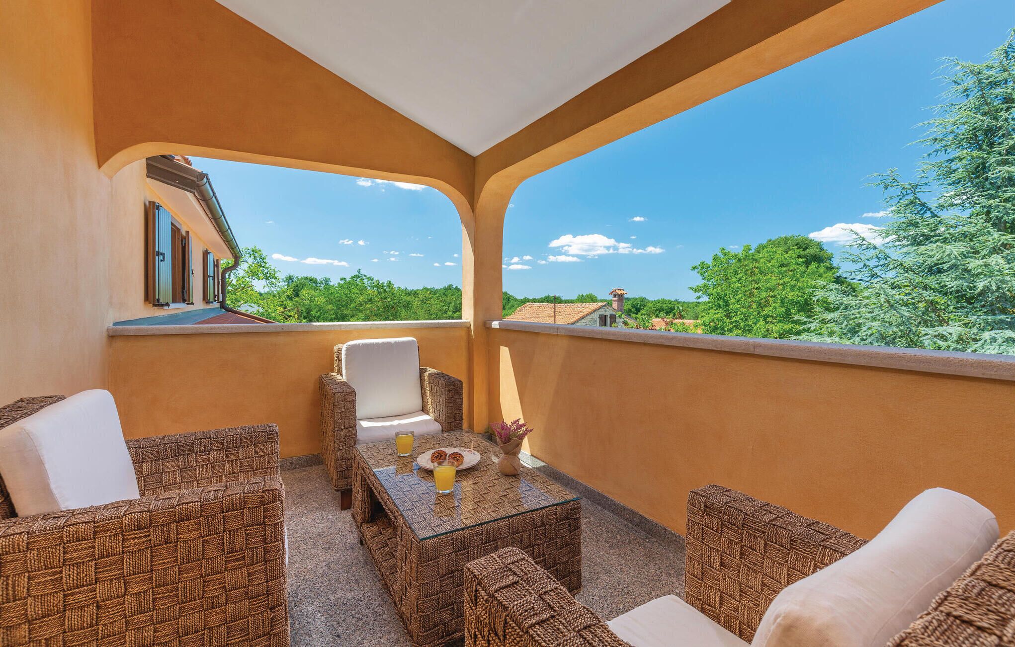 A balcony with wicker furniture, a table with a glass top, and a view of greenery and buildings.