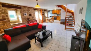 Ein gemütliches Wohnzimmer mit Steinwand, einem schwarzen Sofa mit roten Kissen, einem Couchtisch und einem Fernseher. Im Hintergrund ist eine Küche mit Holzausbau und einer Treppe erkennbar.