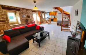 A cozy living room with a stone wall, a black sofa with red cushions, a coffee table, and a TV. A kitchen is visible in the background with wooden cabinets and a staircase. - Stunning home in Plouhinec with kitchen (Plouhinec)