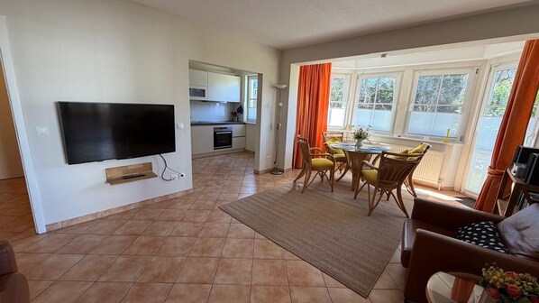 Ein Wohnzimmer mit Flachbildfernseher, ein Essbereich mit Tisch und Stühlen sowie eine Küche im Hintergrund.