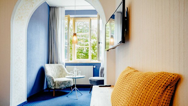 A room with a blue carpet, a white sofa, a small table, two chairs, a window with a view, and a mounted TV. - Standard double room (21m��) - Hotel & Restaurant Alpenrose Bayrischzell (Bayrischzell)