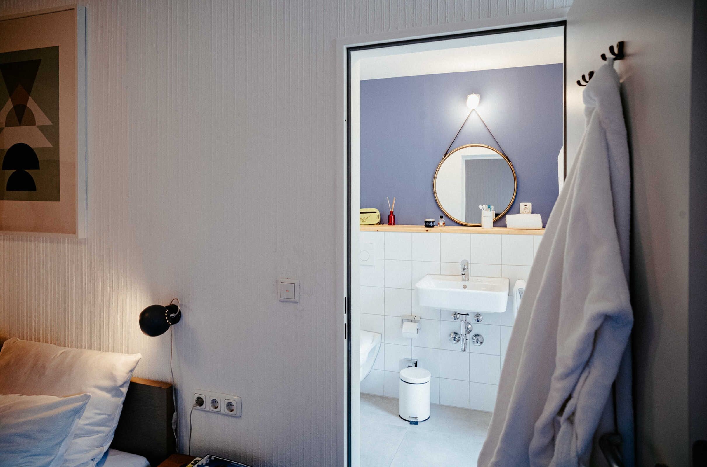 Quarto de hotel com cama, luminária de cabeceira, banheiro com pia, espelho e porta-toalhas.
