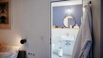 Quarto de hotel com cama, luminária de cabeceira, banheiro com pia, espelho e porta-toalhas.
