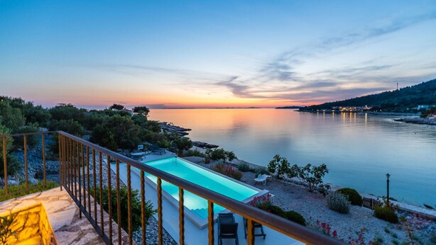 Ein Pool mit Blick auf den Sonnenuntergang über ruhigem Wasser, umgeben von viel Grün und einer Holzterrasse.
