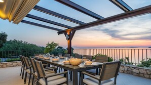 Ein Essbereich im Freien mit Blick auf das Meer bei Sonnenuntergang, mit einem langen Tisch, Stühlen und einer Pergola darüber.