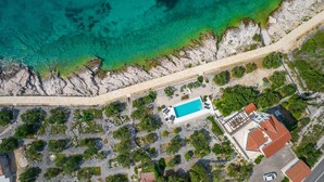Eine Luftaufnahme eines Küsteneigentums mit Schwimmbad, einem gepflasterten Weg entlang des Strandes und einem Gebäude mit rotem Ziegeldach.