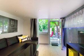 A modern living room with a black sofa, a glass coffee table, and a flat-screen TV. There is a large window with curtains, a radiator, and a view of the outside. - Holiday apartment only 250 m from the sandy beach on the Gold Coast (Siófok)