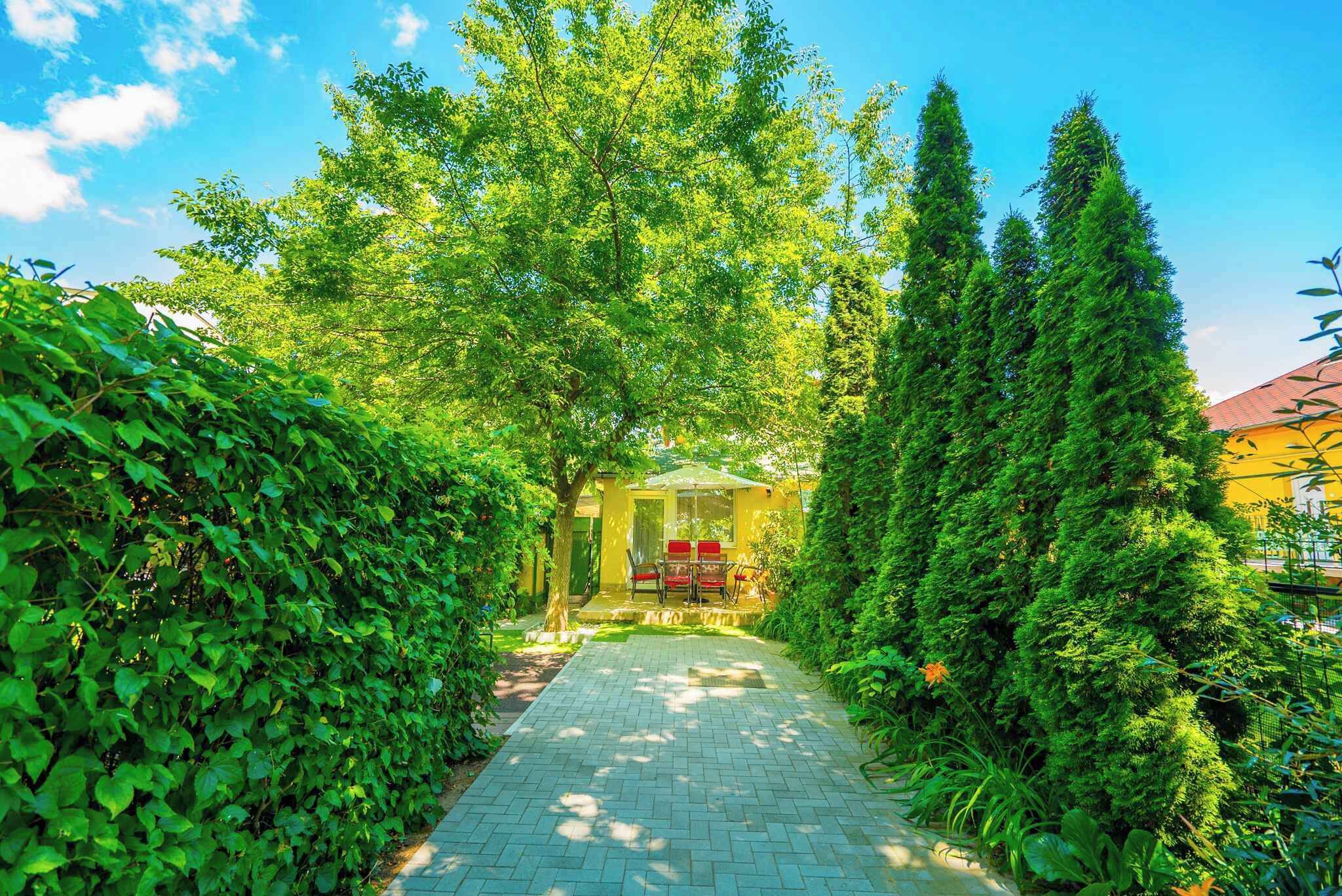 A pathway leading to a yellow gazebo with red chairs, surrounded by lush greenery and tall trees.