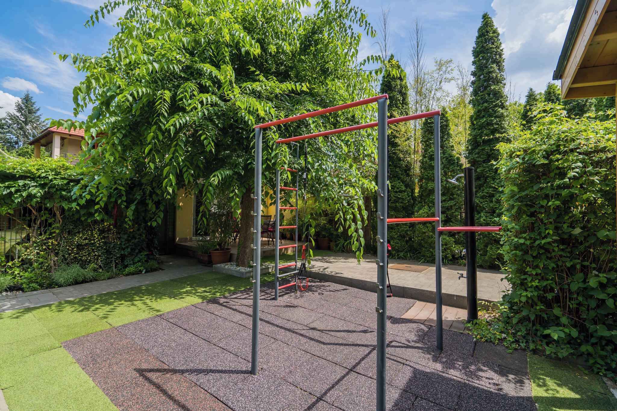 An outdoor gym area with exercise equipment, surrounded by greenery and a building in the background.