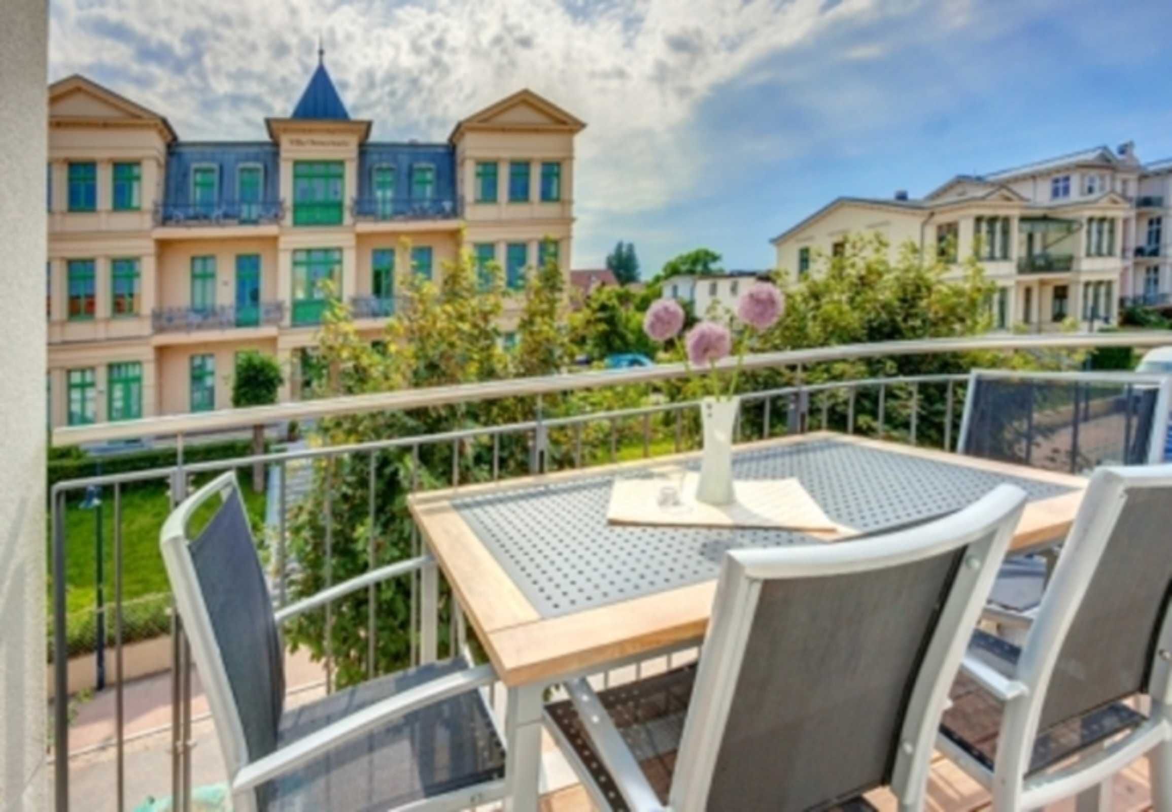 A balcony with a table and chairs, overlooking a row of buildings with multiple stories and balconies.