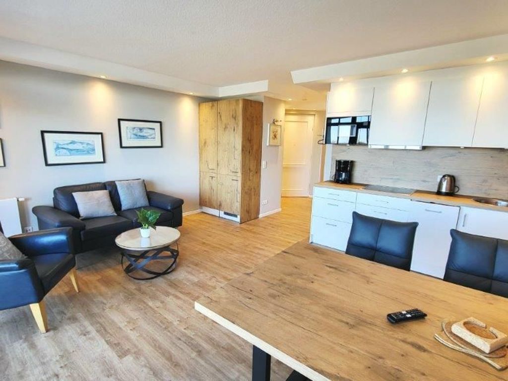 A modern living room with a wooden dining table, black leather sofa, and a coffee table. There is a kitchen area with white cabinets and a black countertop.