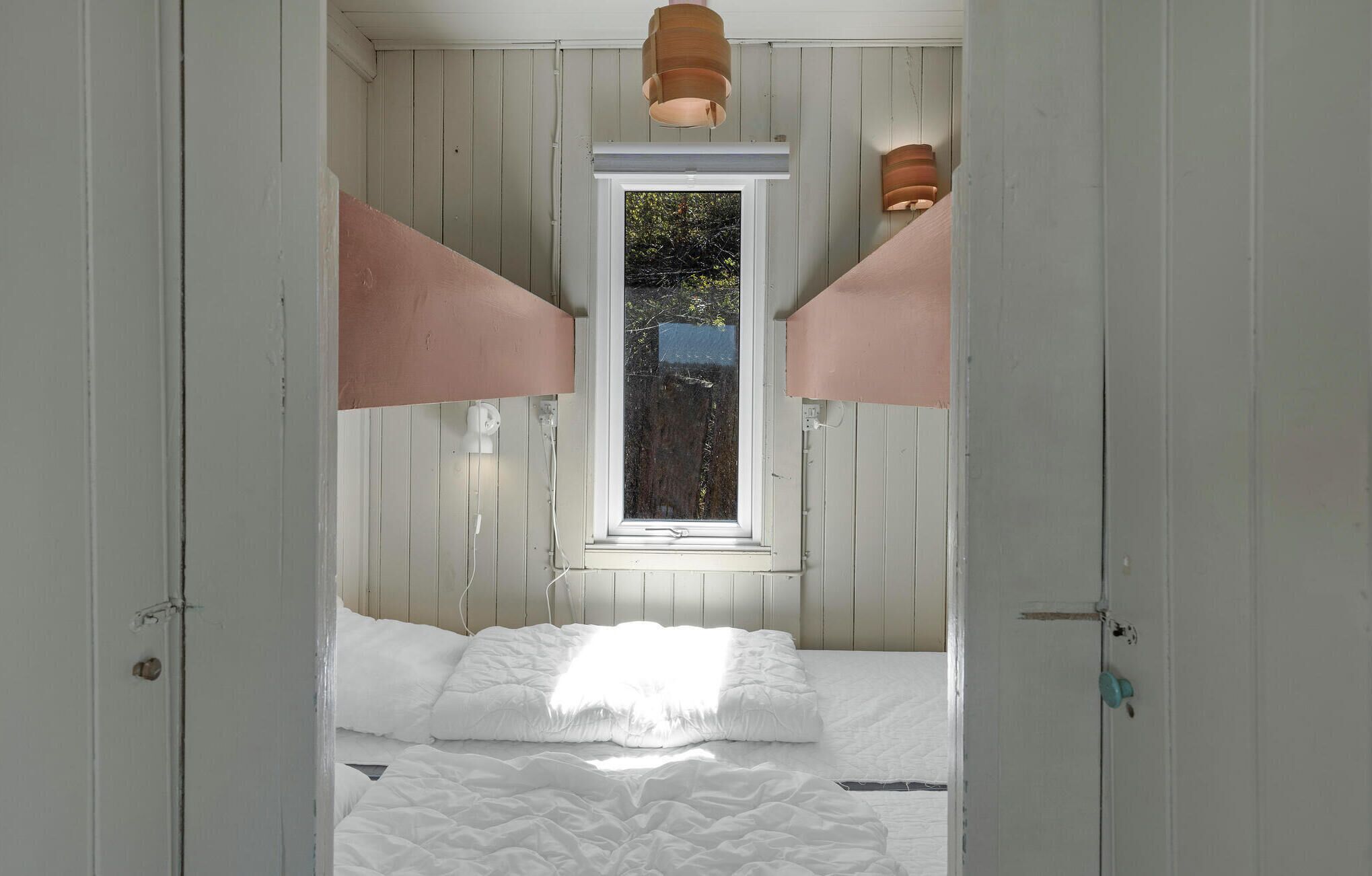 A bedroom with a bed, white walls, wooden paneling, a window, and two wall-mounted lamps.