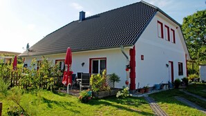 Una casa blanca con techo negro, detalles rojos y un jardín con plantas en macetas y un paraguas rojo.