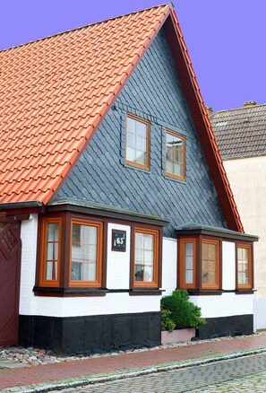 A building with a steeply pitched roof, white walls, and a red-tiled roof. There are three windows with wooden frames and a small green bush in front.