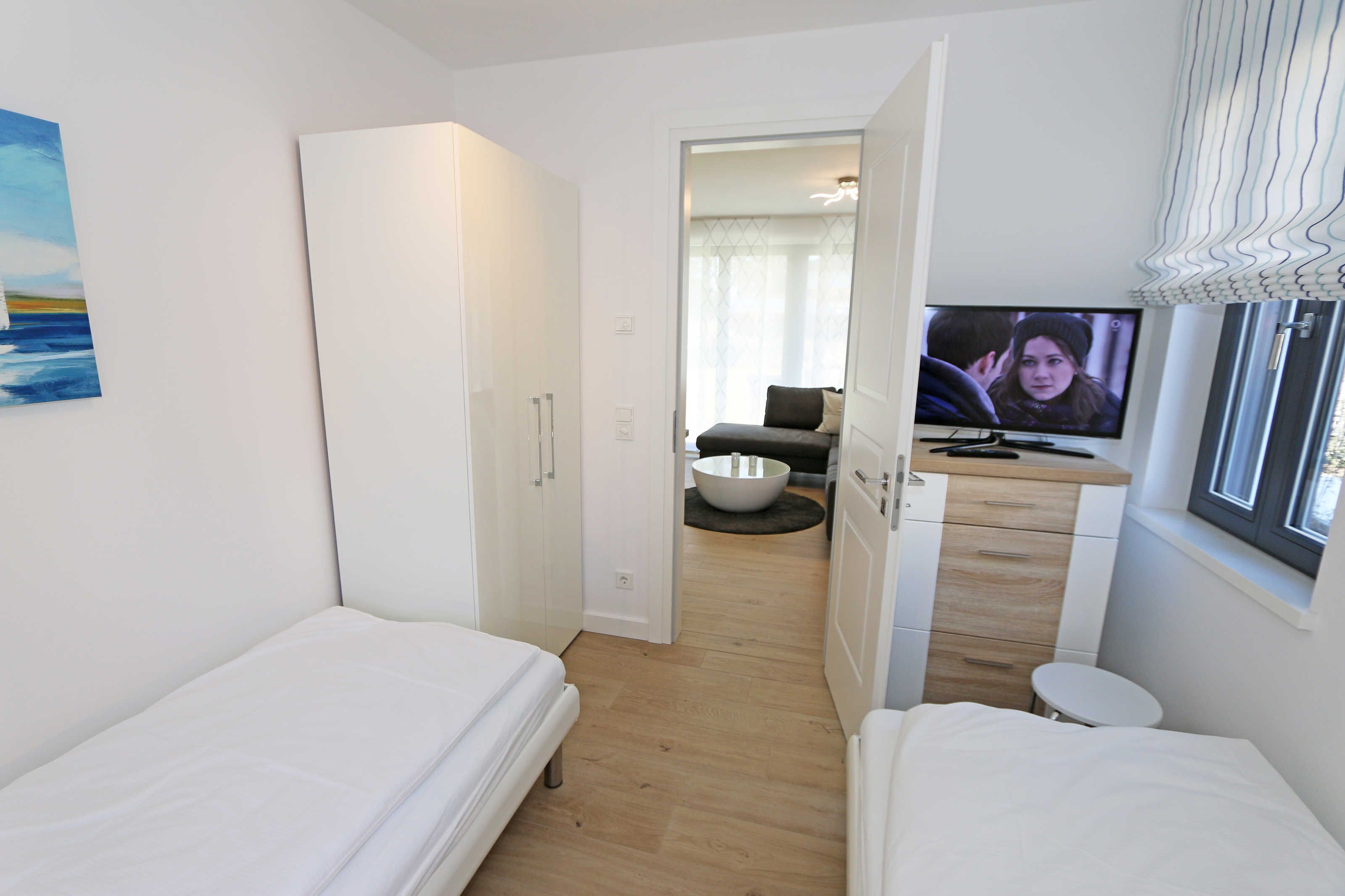 A modern hotel room with two single beds, a TV, a small bathroom, and a view of the living area.