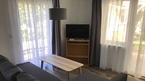 A living room with a sofa, a coffee table, a TV on a stand, and a window with curtains. - Apartment Type 1/2 + 1 ZuB - Meutzner Trassenheide, "Villa Seute Deern" (Trassenheide)