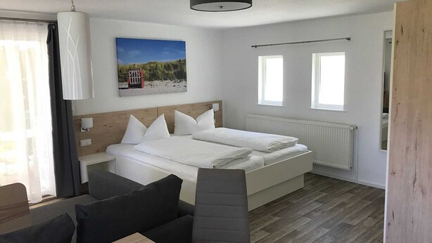 A modern bedroom with a wooden headboard, a large bed with white bedding, a wooden floor, and a view of the beach through a window. - Apartment Type 1/2 + 1 ZuB - Meutzner Trassenheide, "Villa Seute Deern" (Trassenheide)