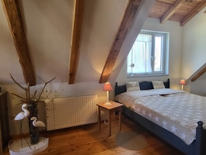 A bedroom with a bed, bedside lamps, a radiator, a window with curtains, and wooden beams on the ceiling. - Blue Water 2, Germany (Wustrow)