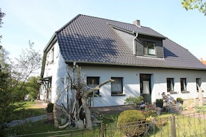A white house with a grey tiled roof, a visible chimney, and a small garden with potted plants and a tree. - Blue Water 2, Germany (Wustrow)