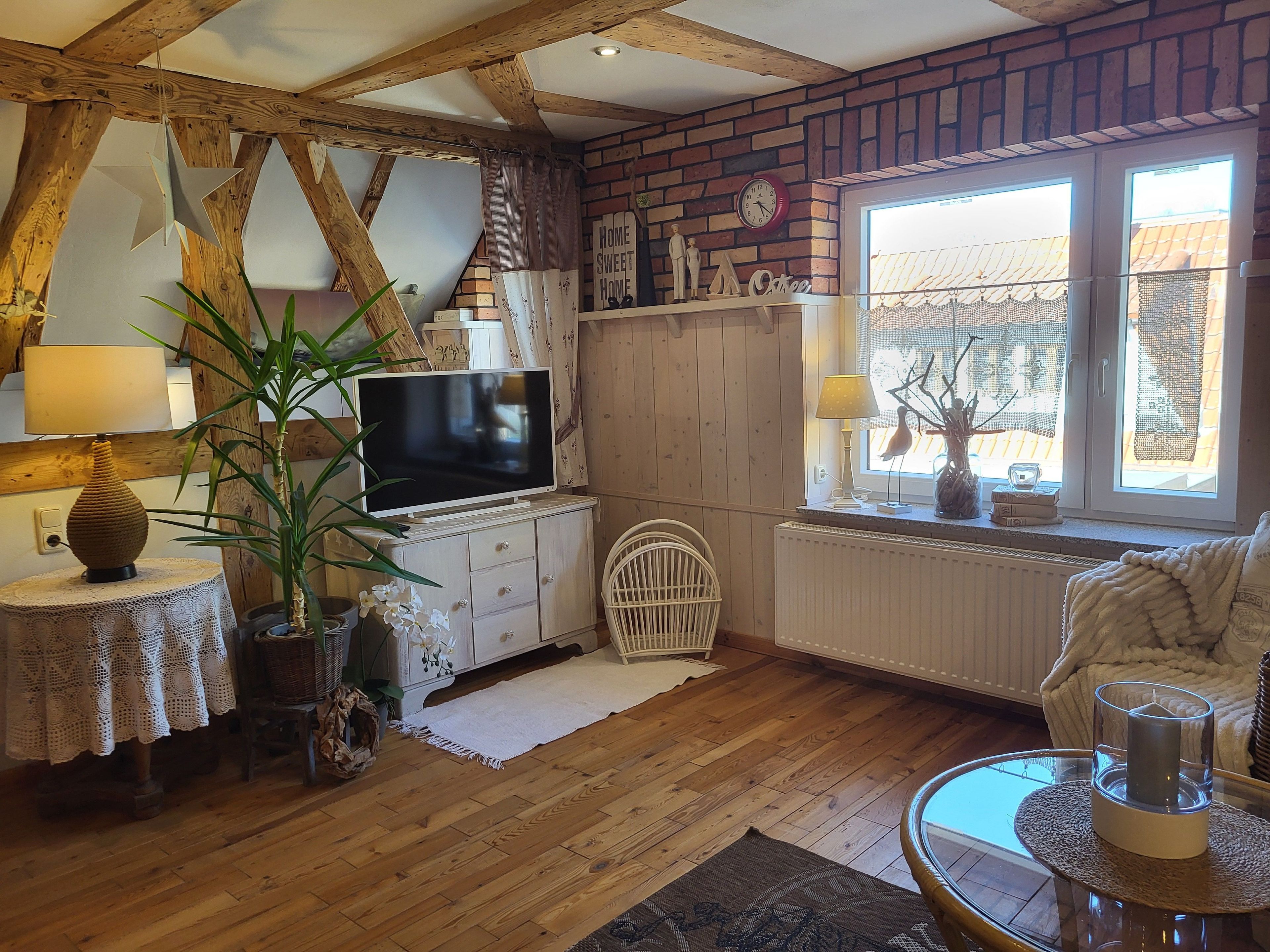 Una sala de estar acogedora con vigas de madera a la vista, un televisor sobre un mueble de madera, un sillón de mimbre, un sofá blanco y una ventana con vistas al exterior.