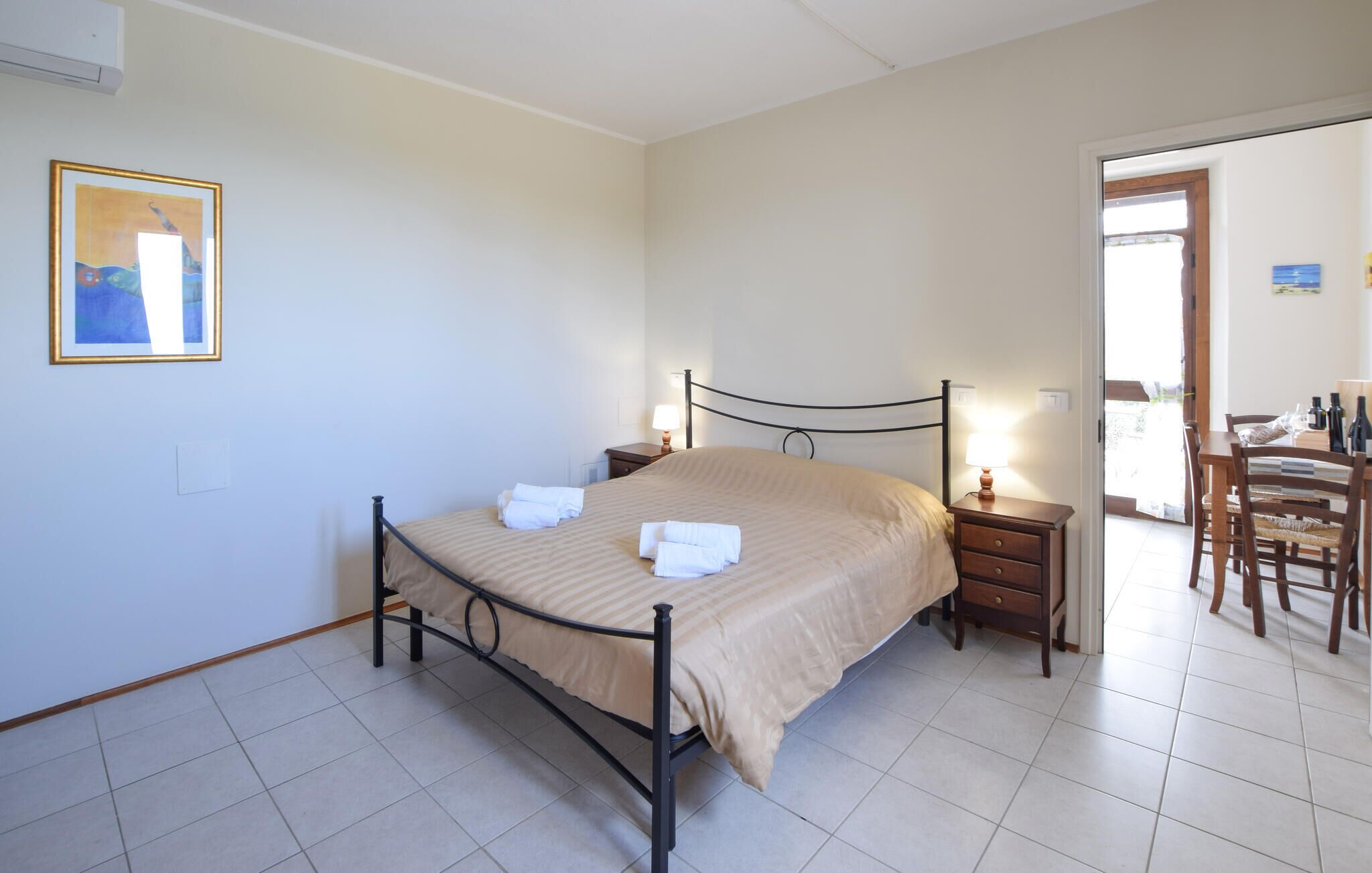 Una cama bien hecha con una colcha a rayas y dos toallas dobladas, una mesita de noche con una lámpara, y una silla con una mesa lista para comer.