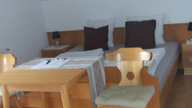 A hotel room with a wooden bed, a wooden nightstand with a lamp, a wooden table, and a chair. - Dachsteinblick - Organic Farm Mondsee Family Schafleitner-Kroiß (3 Flowers) (Guggenberg)