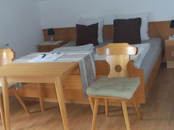 Habitación de hotel con una cama de madera, una mesita de noche de madera con lámpara, una mesa de madera y una silla.