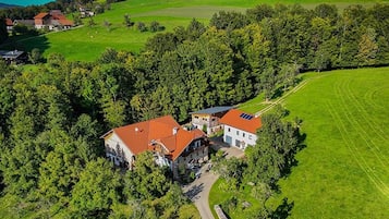 Vue aérienne d’une propriété rurale comprenant deux bâtiments principaux, entourée d’une végétation luxuriante et surplombée d’un ciel dégagé.