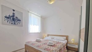 A bedroom with a wooden bed, white bedding, a window with sheer curtains, a wall-mounted light fixture, and a bedside table with a lamp. - Holiday apartment newly furnished for 2-4 pers. with air conditioning, WLAN and car park (Baška)